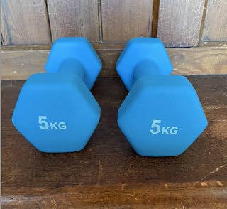 Two dumbbells, sitting on a bench. They're a turquoise blue and say 5kg on the end of each one.