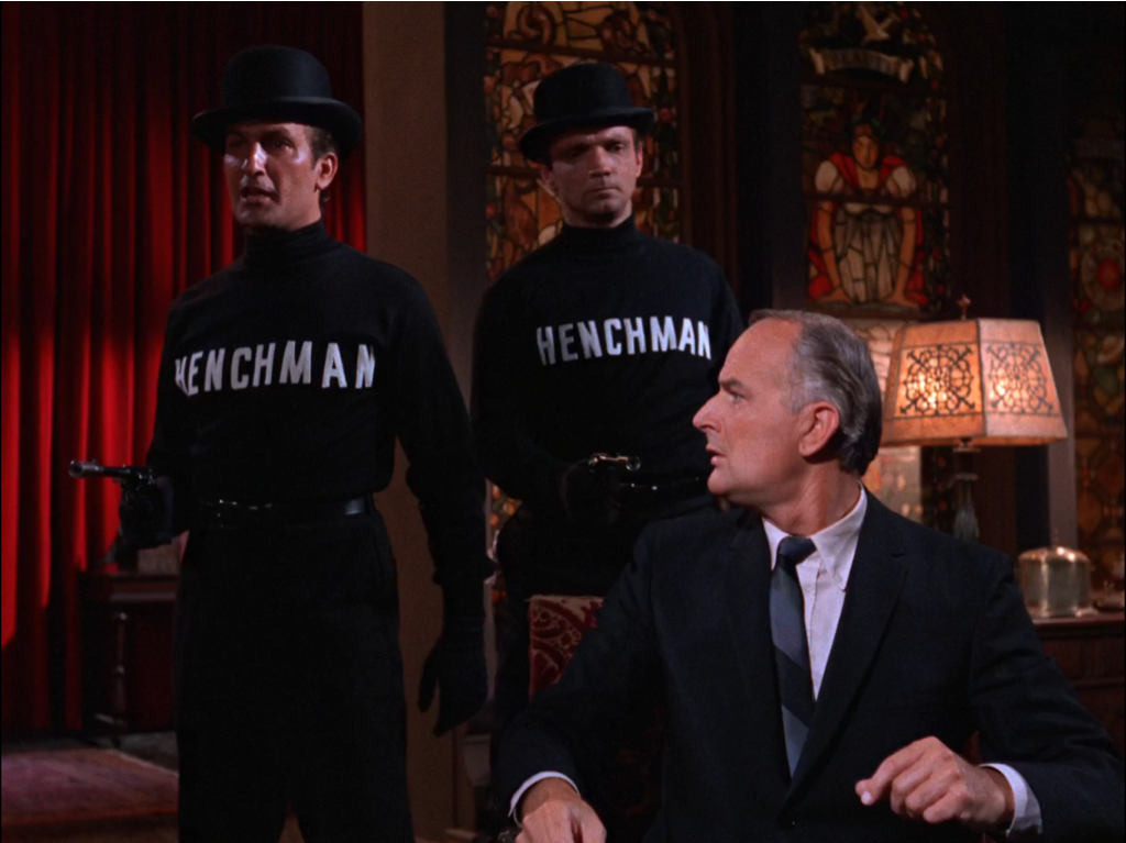 Still from 1960's Batman TV show - a guy in a suit with two guys behind him wearing black turtlenecks that say "HENCHMAN" in white block letters