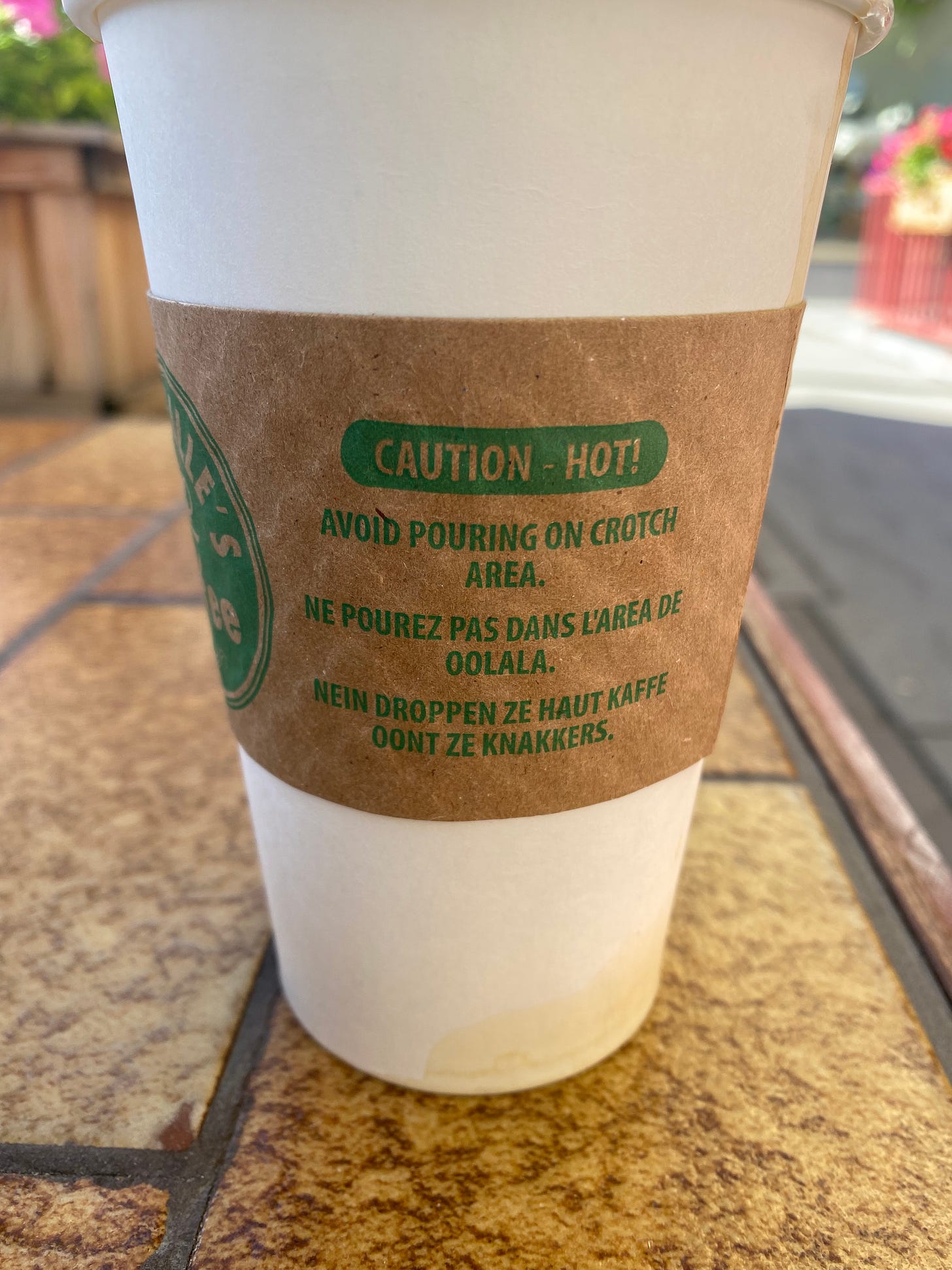 A takeaway coffee cup with a paper sleeve that reads 'caution - hot!' with jokes in several languages below it about not pouring it onto your crotch.