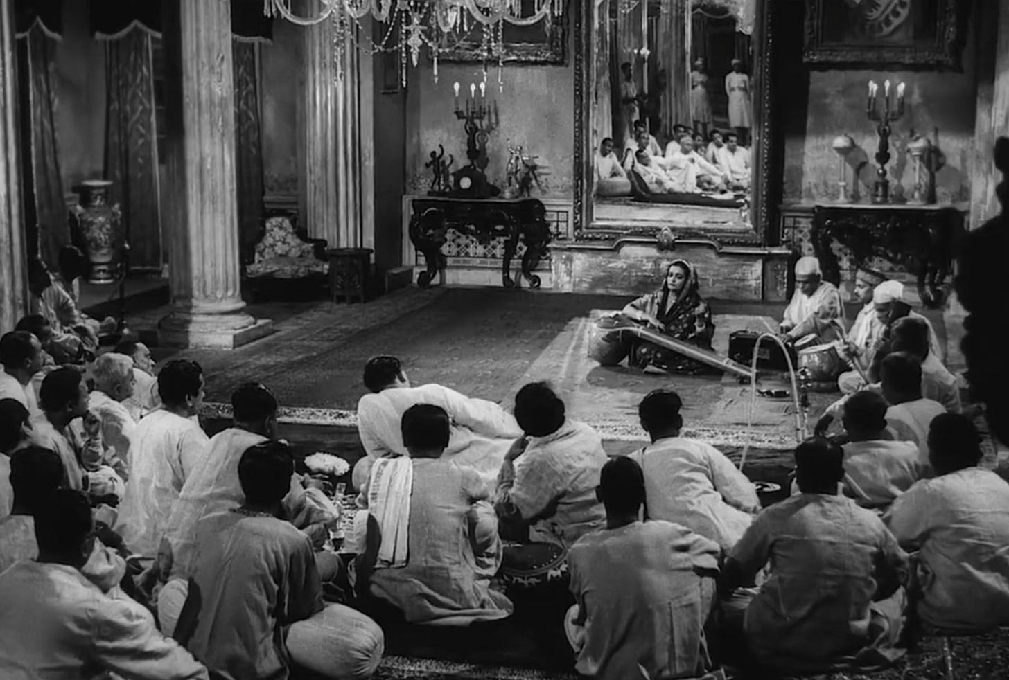 A woman plays a traditional Indian string instrument with a large male audience in a lavishly decorated room.