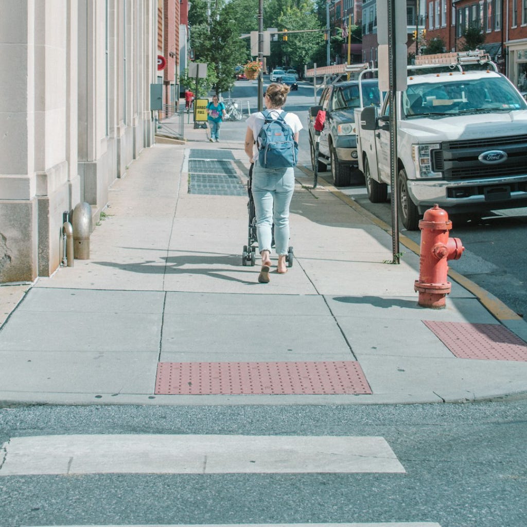 A woman pushing a stroller walks away from the camera, a curb-cut corner is in the foreground.