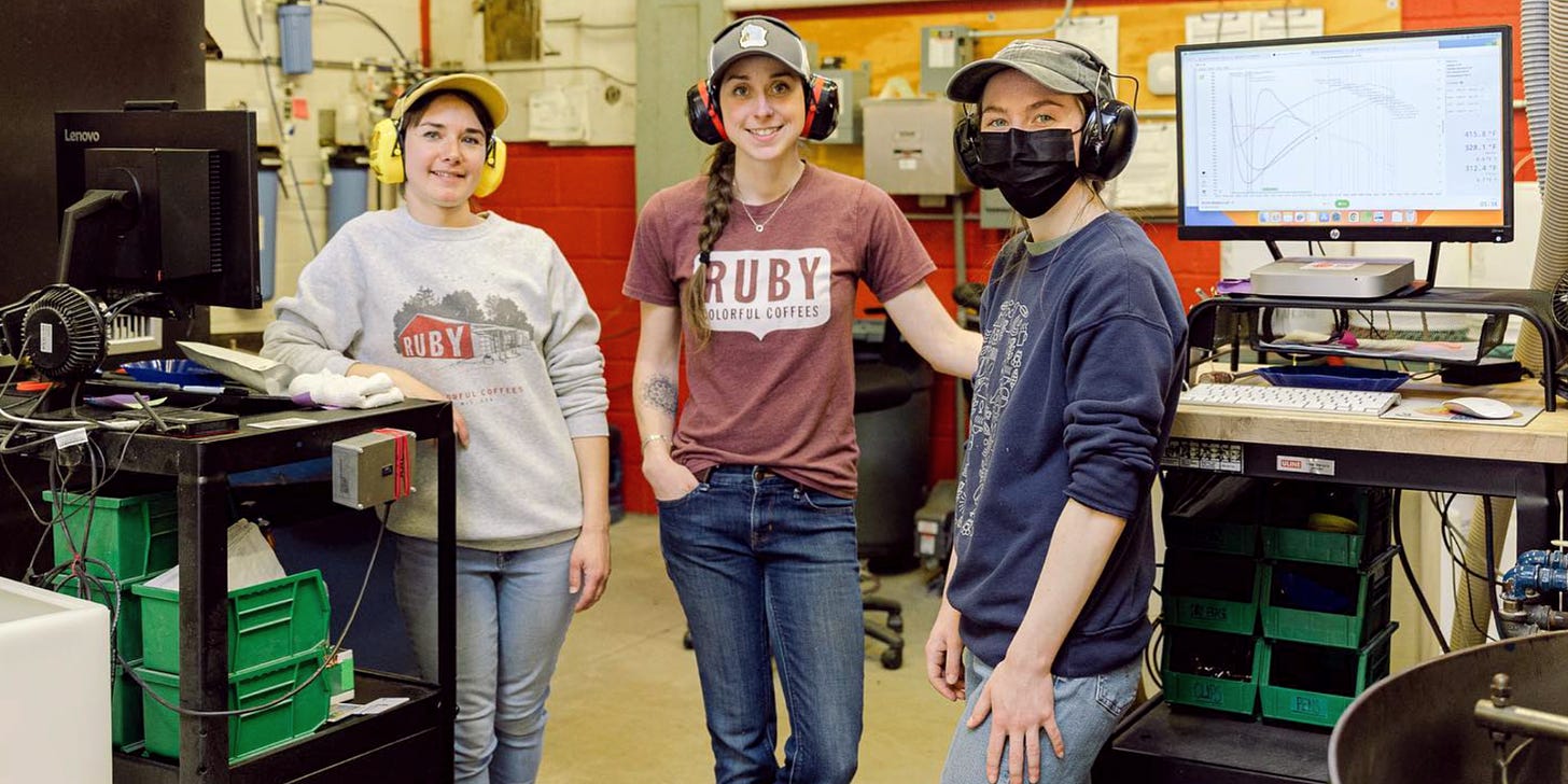 Three people wearing ear protection, baseball caps, jeans, and Ruby Coffee shirts smile at the camera from inside a coffee manufacturing facility.