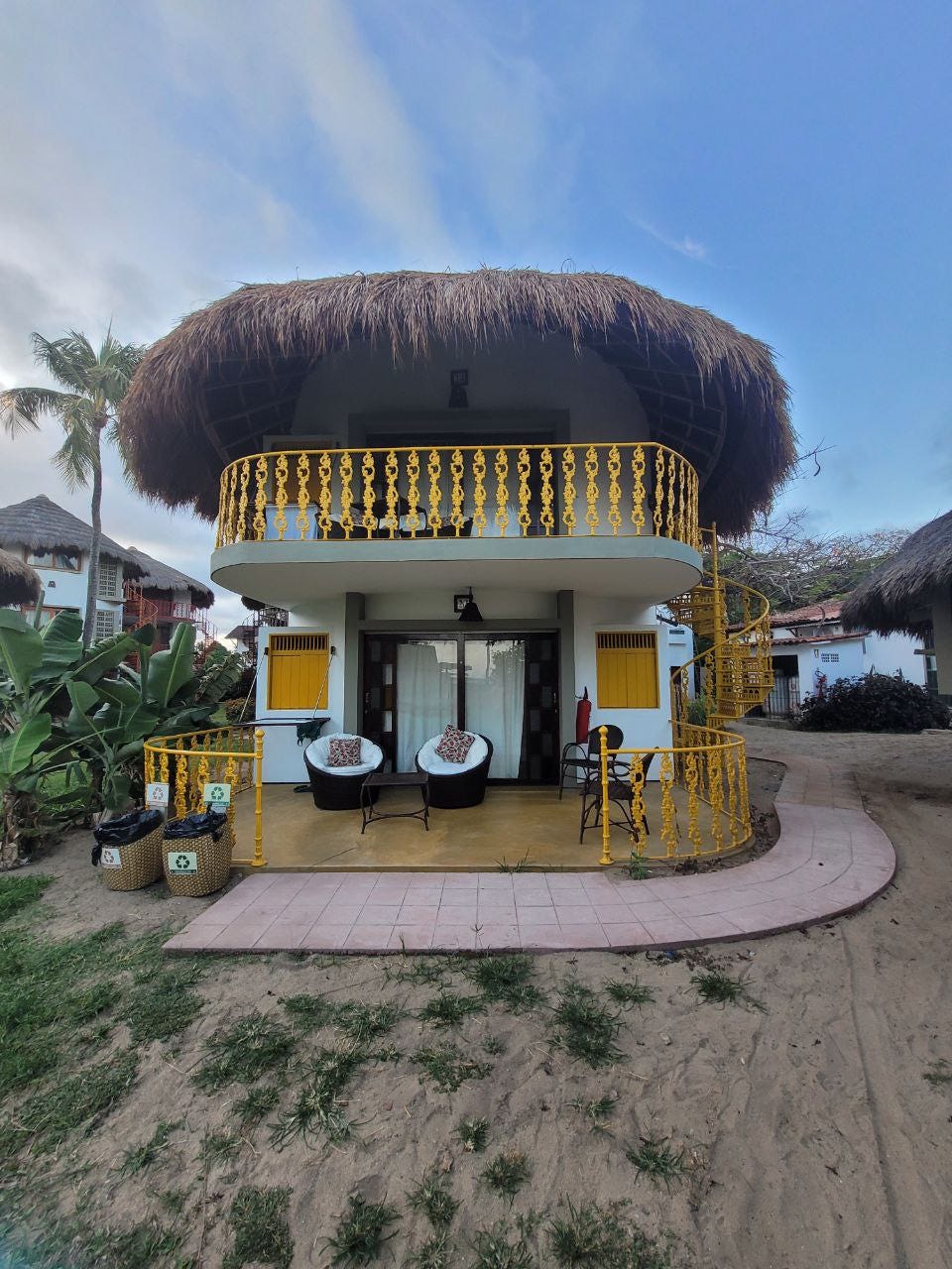 Numa areia de praia há um bangalô com teto de palha e grades amarelas. Ele tem dois andares, no de cima dá pra ver uma varanda e no de baixo também, com poltronas e cadeiras de frente a uma porta de vidros. Dá pra ver no fundo outros bangalôs similares.