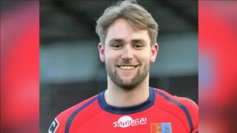 Chester RUFC Jon Devereux is seen from his shoulders up wearing a red rugby uniform. He is smiling.