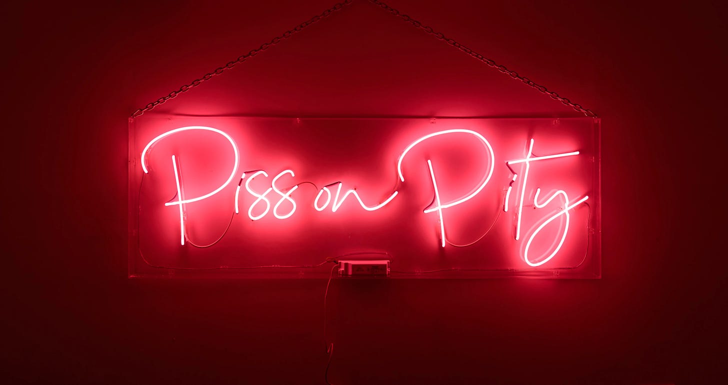 Neon glass tube lights surrounded by a Perspex box with electric plug. The sign says Piss on Pity.