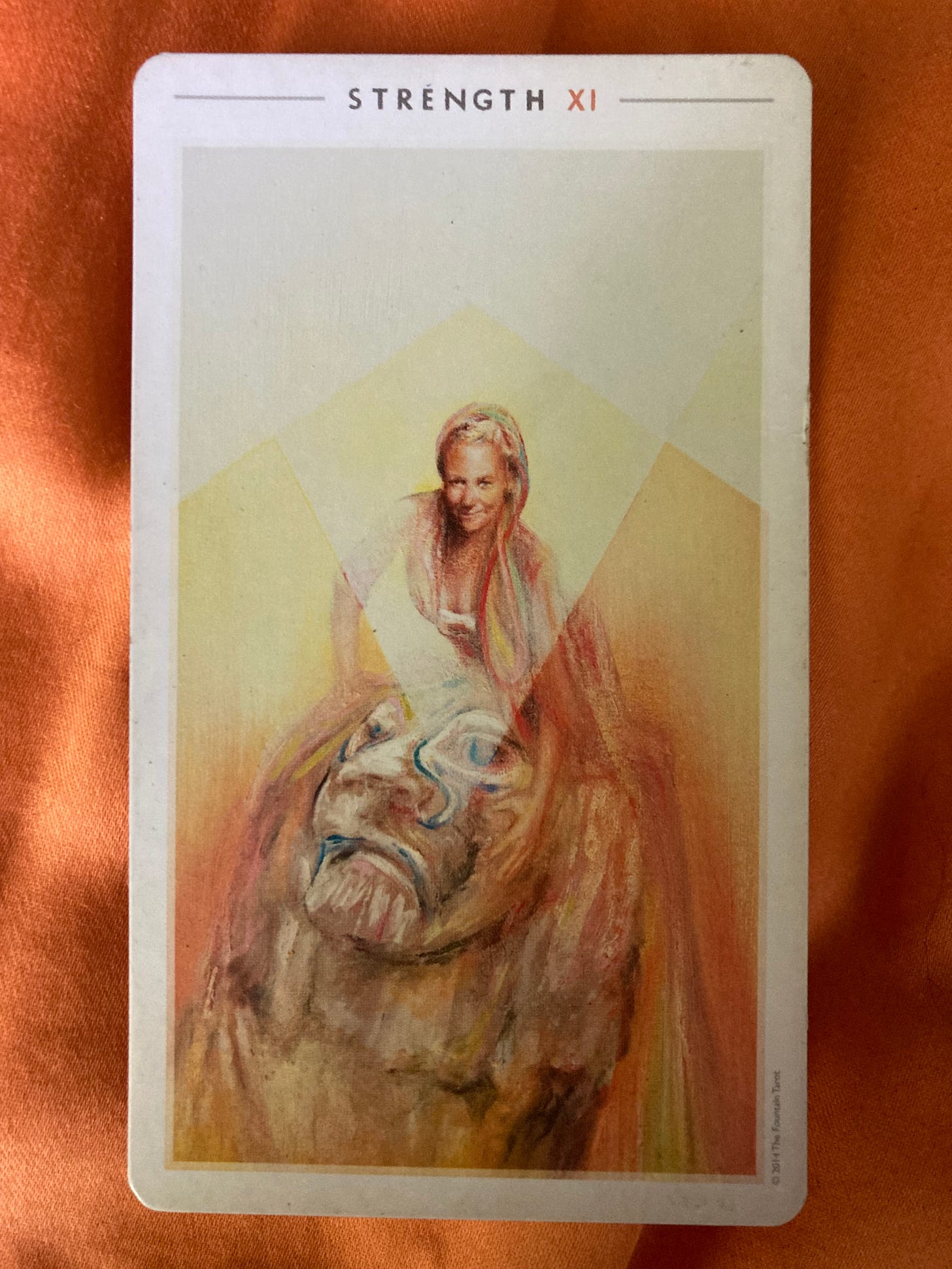 The Strength tarot card lays on an orange background. The card shows a drawing of a young woman sitting on the back of a lion