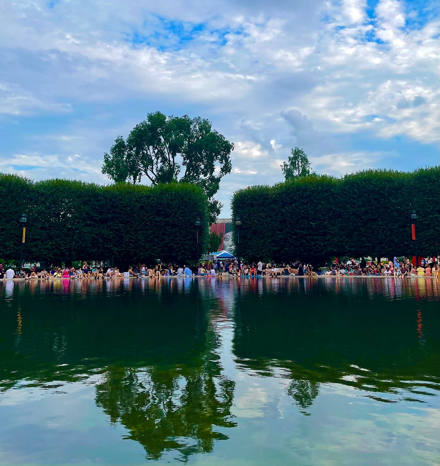 National Sculpture Garden, Washington DC, July 2022, a blue sky and green water reflecting the blue sky, people are sitting around the water, and big bushes are behind them. In between the bushes there is a painting of a women peering out