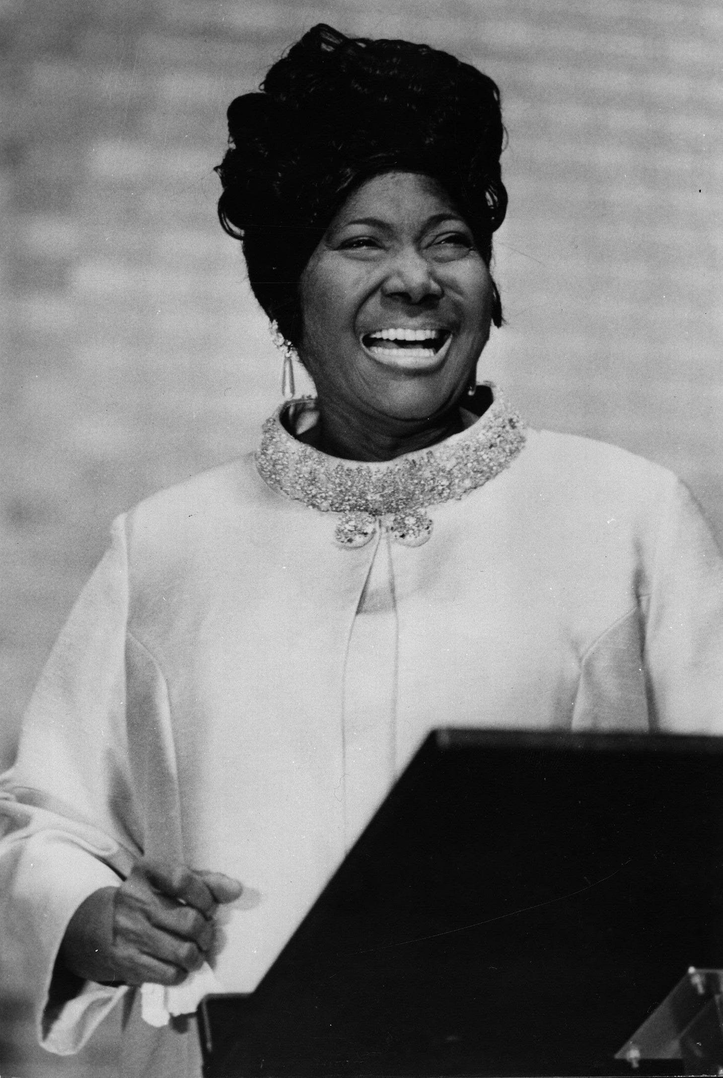 Black and white photo of Mahalia Jackson at a lectern dressed in white and smiling broadly