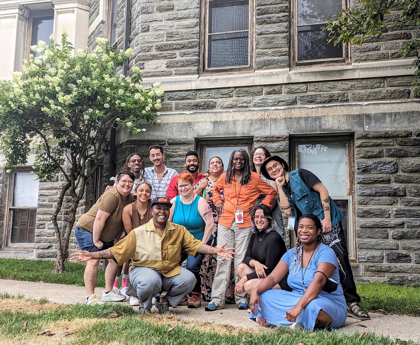 The YA Fiction cohort, a diverse group of queer writers smiling and posing in front of a grey brick building
