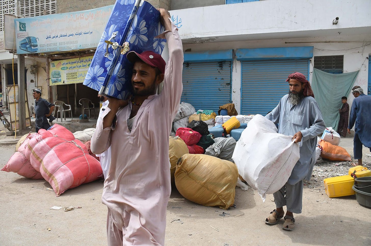Pictured are two Afghan refugees carrying packaged, one carries a blue luggage over his shoulder and another a white sack. They are outside of a building to leave the Pakistani port city of Karachi and return to Afghanistan following the Pakistani government's decision to deport those illegally staying in the country, in Karachi, Pakistan on October 06, 2023.