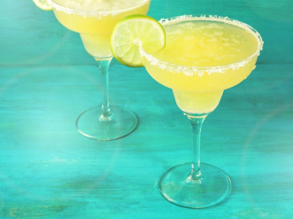 2 bright yellow margaritas in salt-rimmed glasses with lime garnishes against a teal background