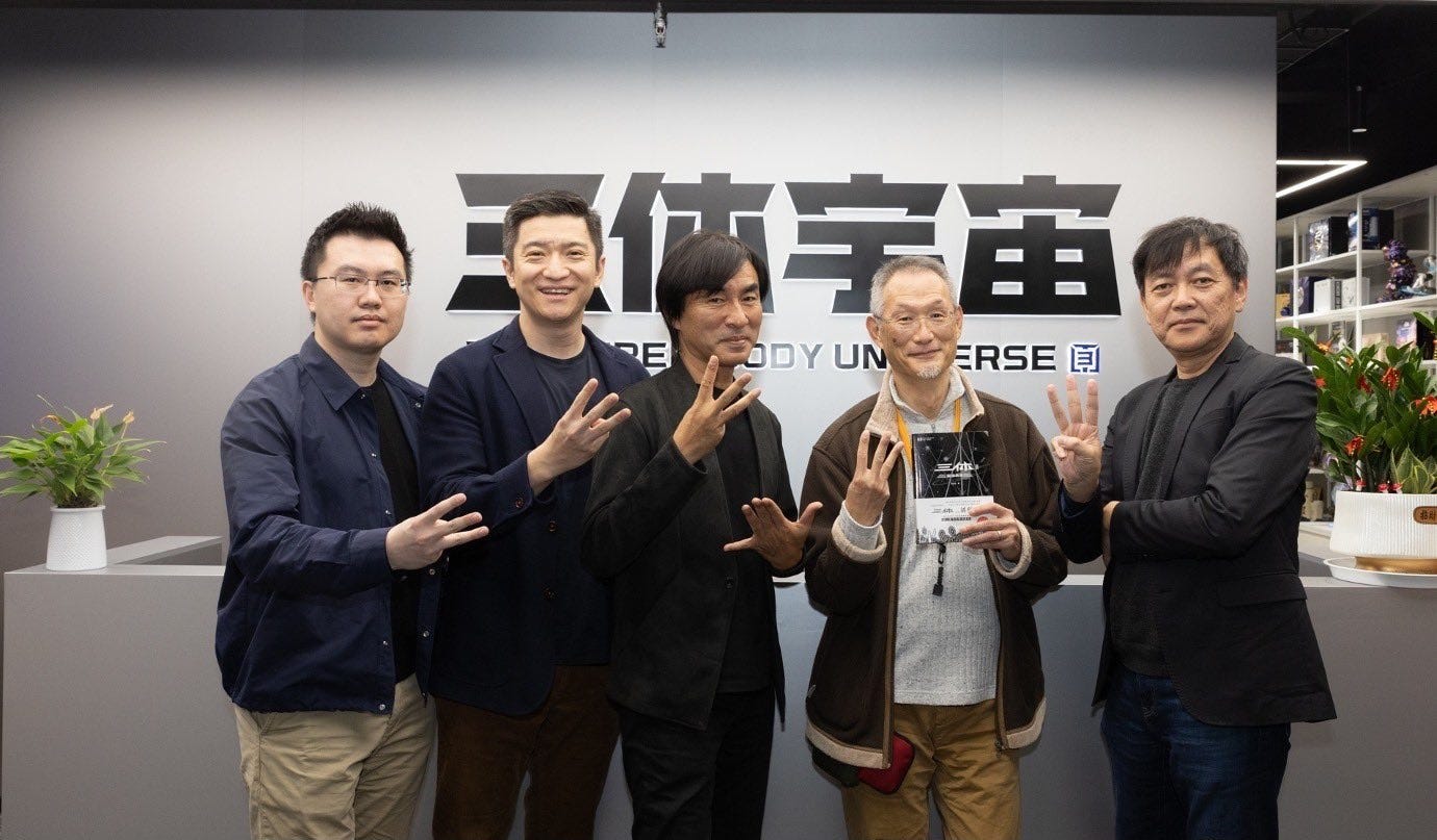 Photograph of five men standing next to each other in an office facing the camera. Each is holding up the three middle fingers of his right hand. Behind them are four Chinese characters and the words "The Three-Body Universe" displayed on the wall.