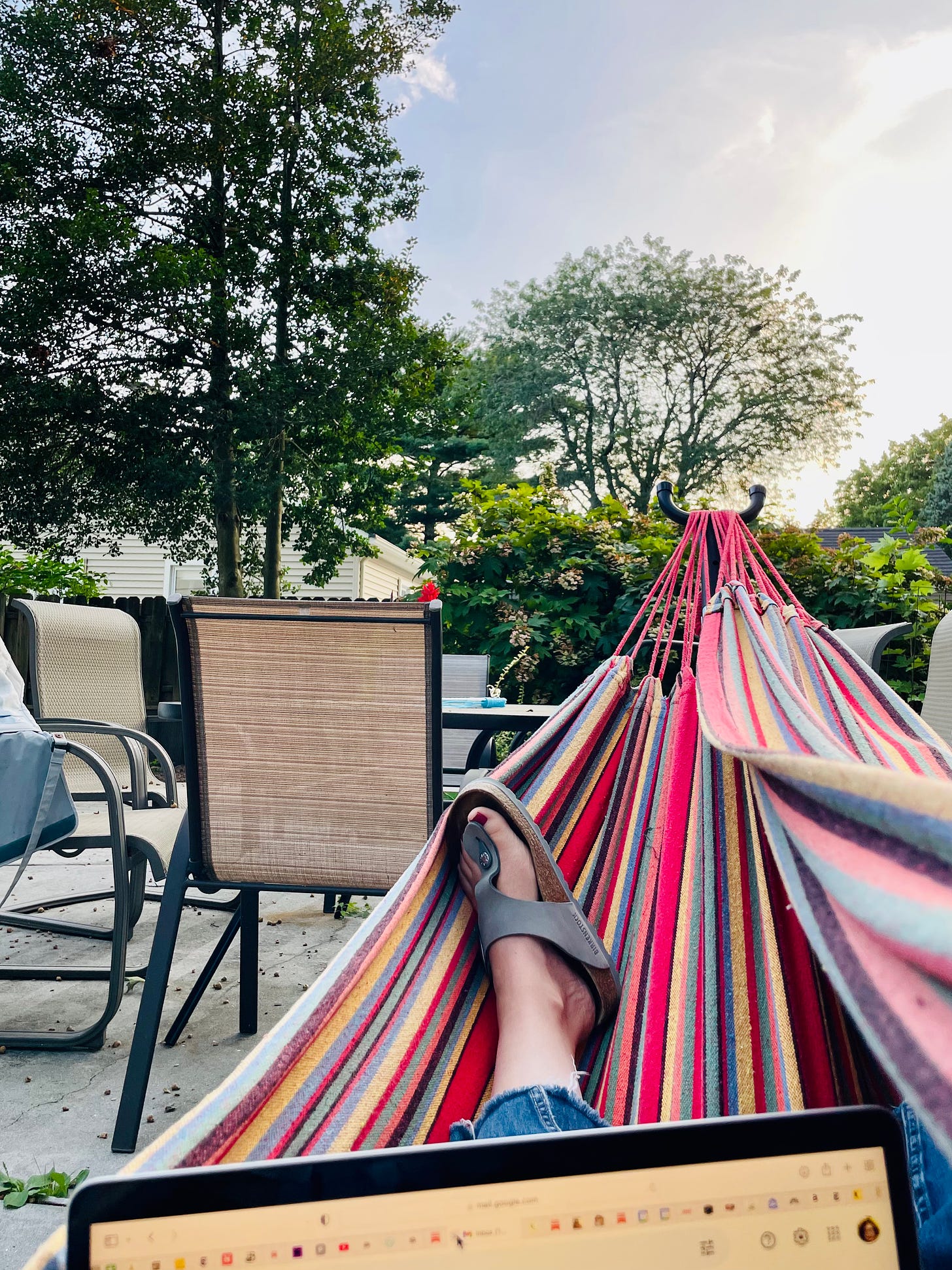 View from a red, yellow, blue, and black striped hammock: A sandaled foot on the hammock, the very top of a laptop, outdoor table and chairs in the background, and trees.