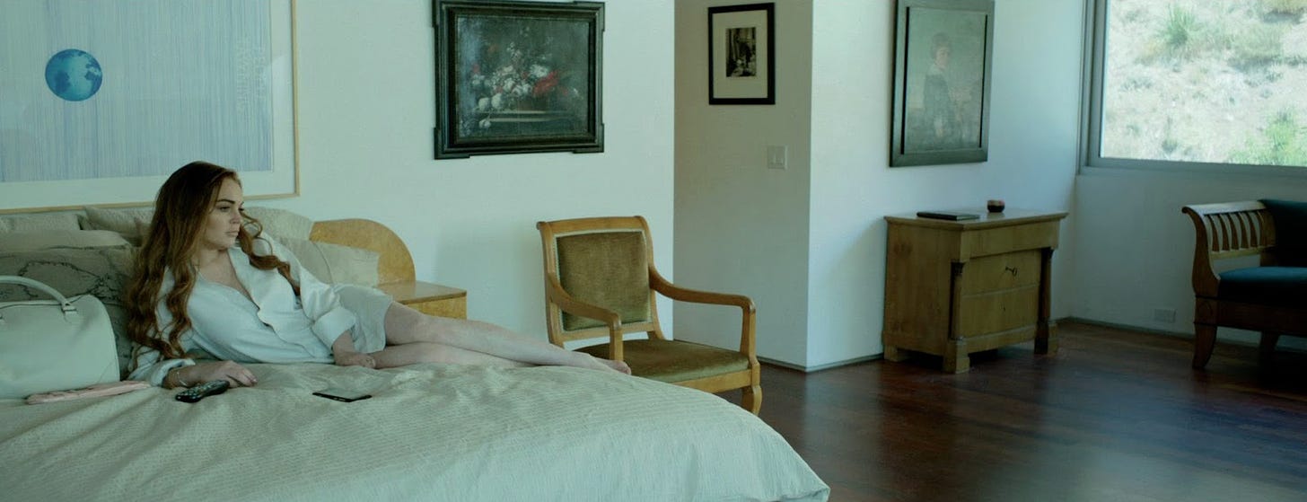 Film still from The Canyons. Lindsay Lohan lounges on a bed in a room, wearing a dress shirt. The room feels cold and empty.