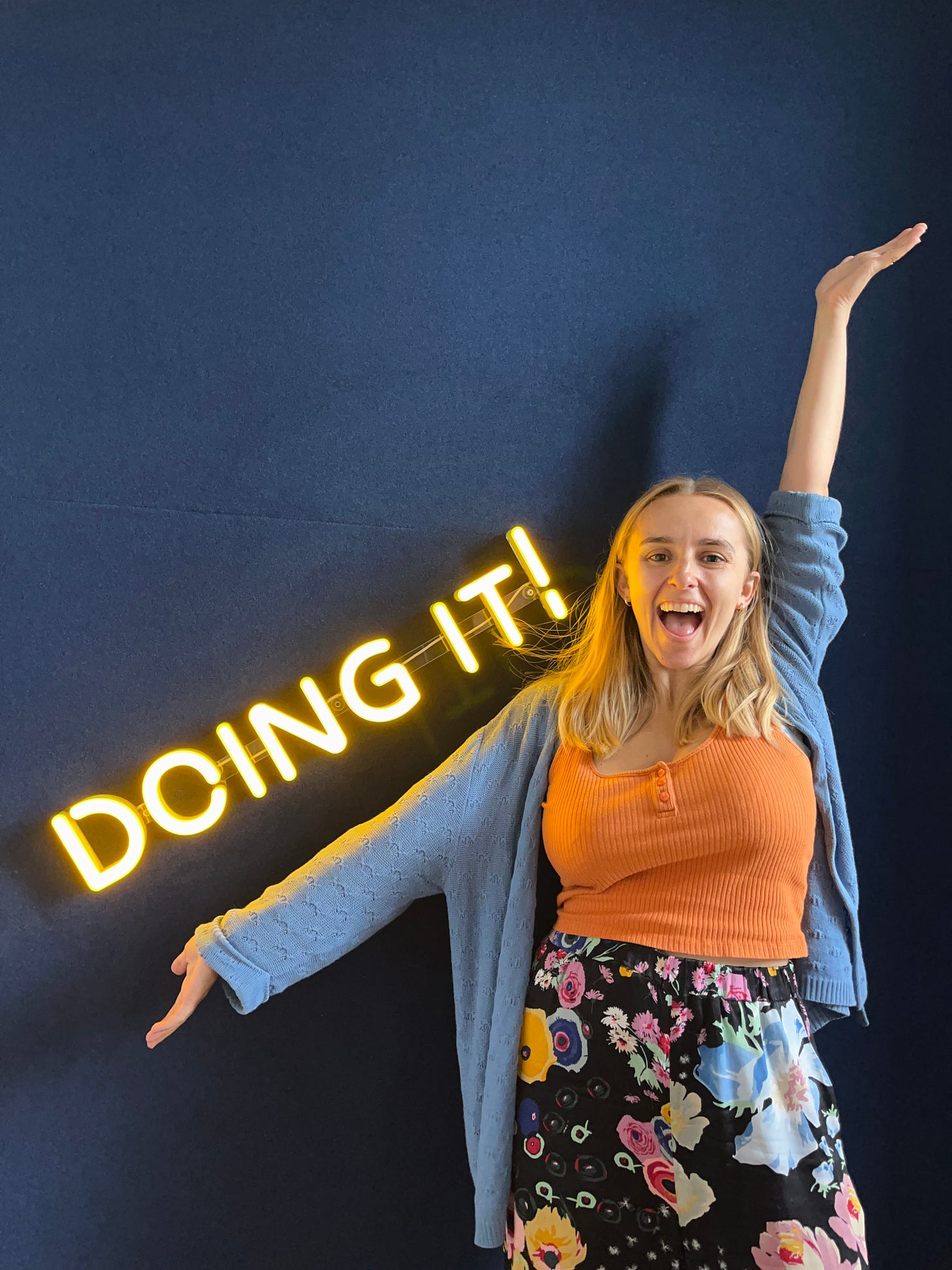 Hannah with her arms out in a "ta-da" pose next to the Doing It neon sign in the Doing It studio