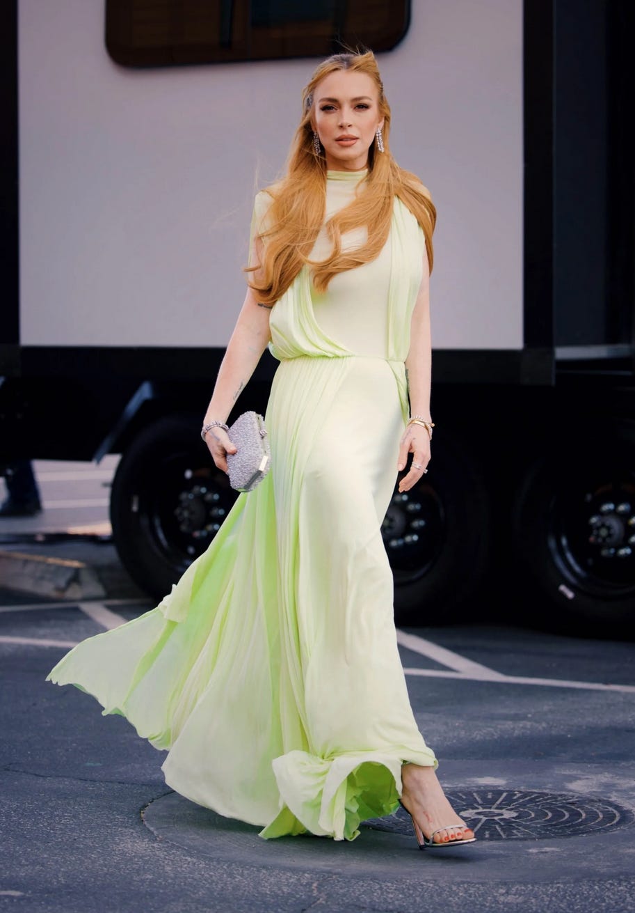 Lindsay Lohan walks confidently on a street while wearing a flowing, light green gown. The dress features a high neckline and drapes elegantly, with soft, billowing fabric that moves as she strides. Her long red hair is styled loosely, cascading over her shoulders. She accessorizes with sparkling earrings, a silver bracelet, and a metallic clutch bag, completing the look with strappy heels. The background includes a black vehicle and muted tones, allowing her vibrant ensemble to stand out.