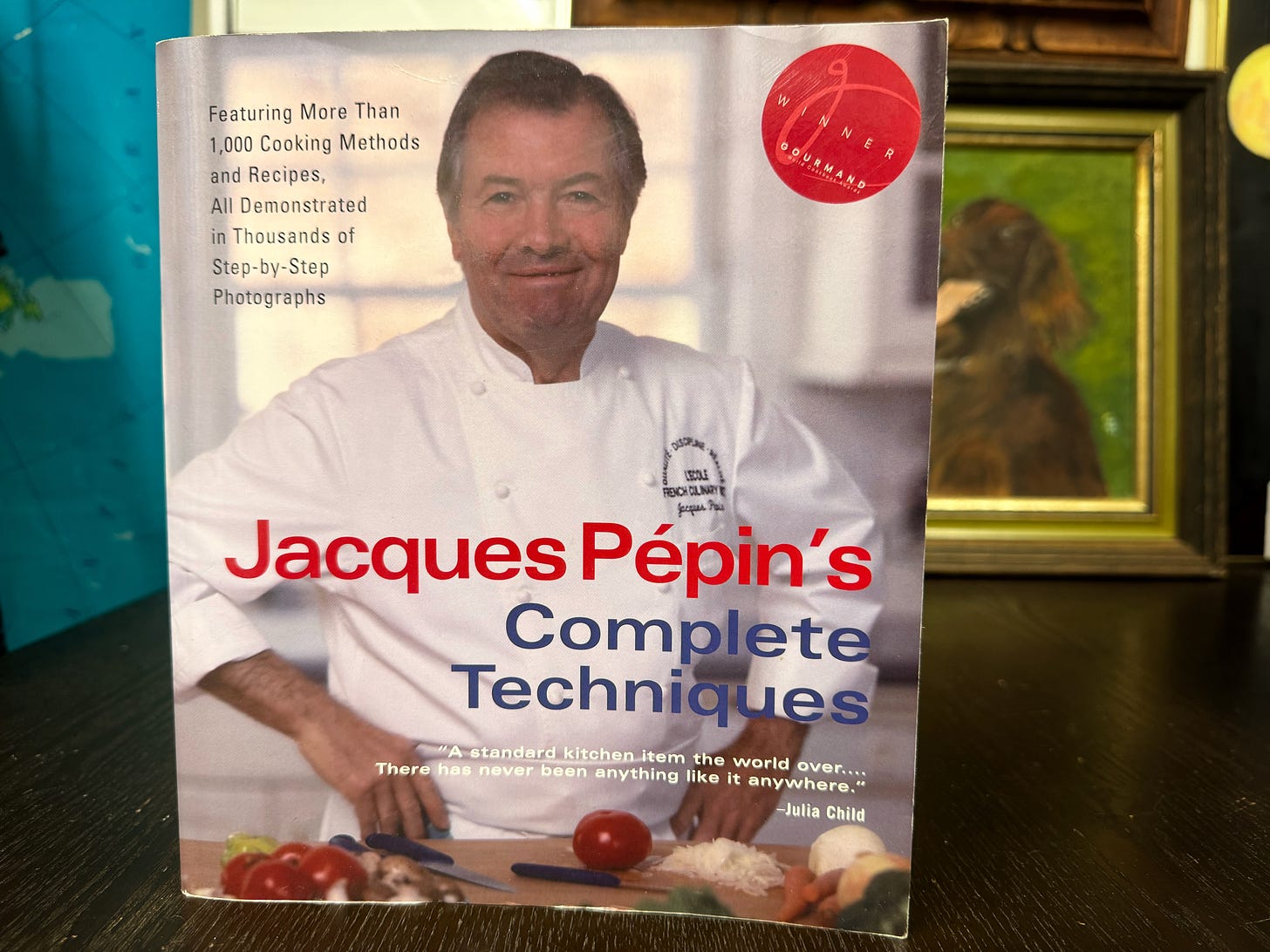 A copy of Jacques Pepin's Complete Techniques on a table. The cover reads, "Featuring more than 1,000 cooking methods and recipes. All demonstrated in thousands of step-by-step photographs." There is a blurb from Julia Child that reads, "A standard kitchen item the world over... There has never been anything like it anywhere."