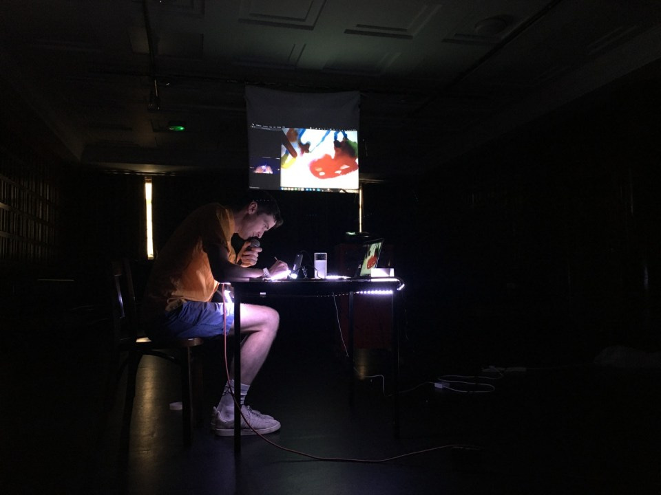 Tim sits at a table, illuminated by lights built into its surface. He is payign close attention to his microscopic painting. Behind him the painting is projected onto a screen. The room is otherwise dark. 