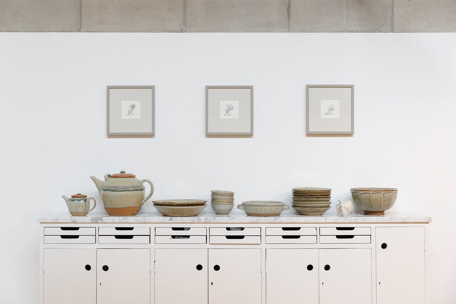 A large sideboard with stoneware pottery piled in a row. Three square picture frames spaced equally above.