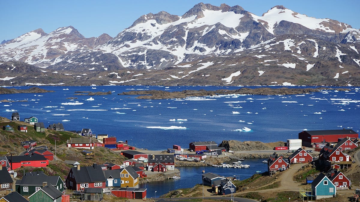 Small houses painted in bright colors dot a hillside in the foreground in front of a fjord with icebergs and, in the background, tall mountains partially covered in snow.