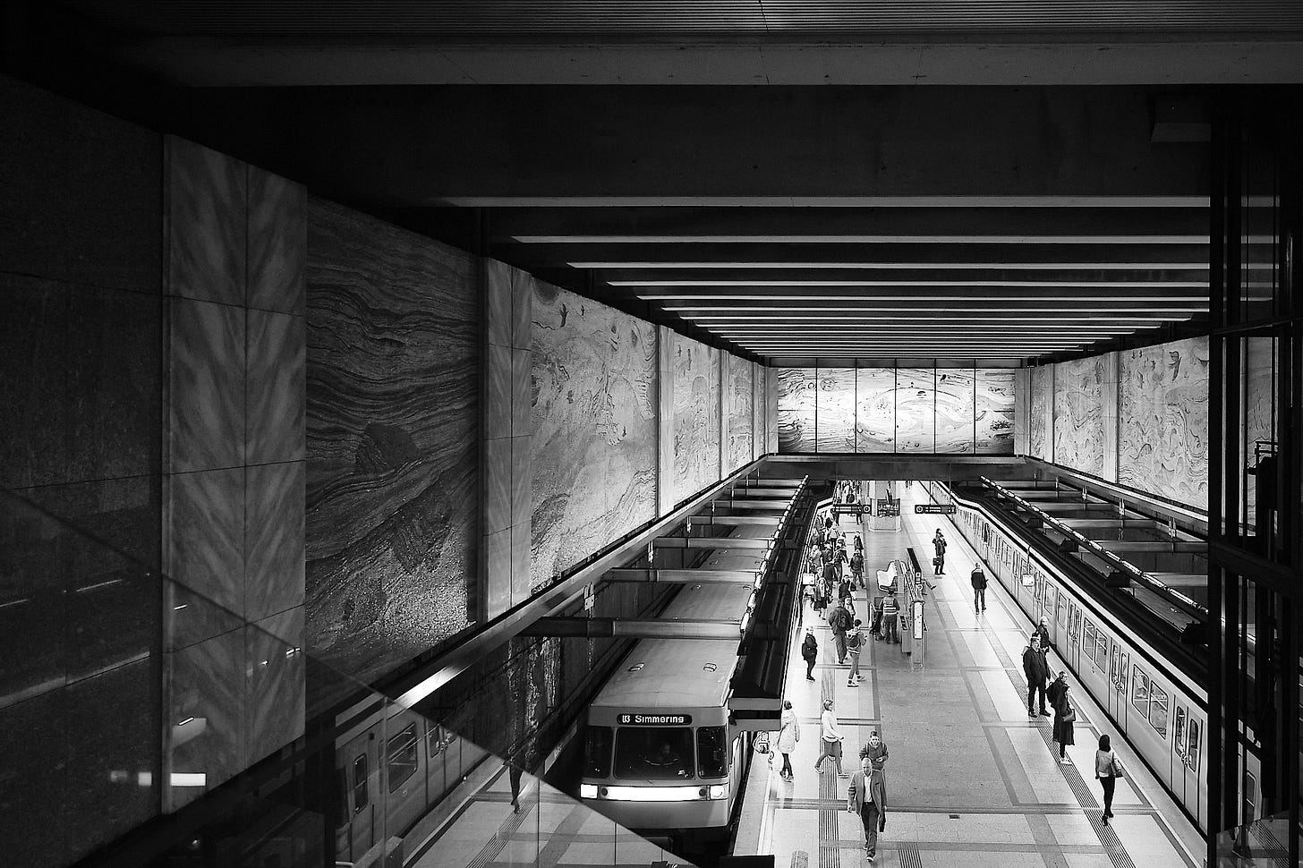 Black and white photograph of the Volkstheater U3 subway station in Vienna. The station features a modern design with sleek lines, a marble-patterned wall on the far side, and a clean platform with passengers waiting. A subway train marked 'U3 Simmering' is stationary on the track closest to the camera, with its doors closed. Overhead strip lighting illuminates the station, and the platform is accessible via escalators and stairs.