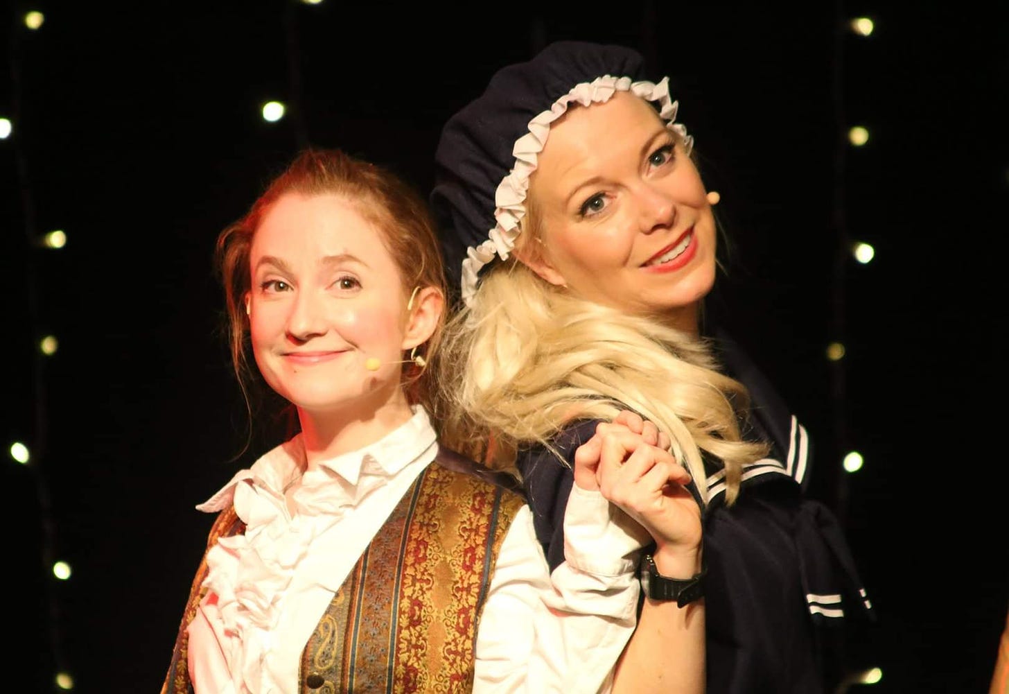Two women stand back to back in panto costumes. Small red head dressed as a panto style lead boy and a woman with blonde hair dressed in a maid outfit