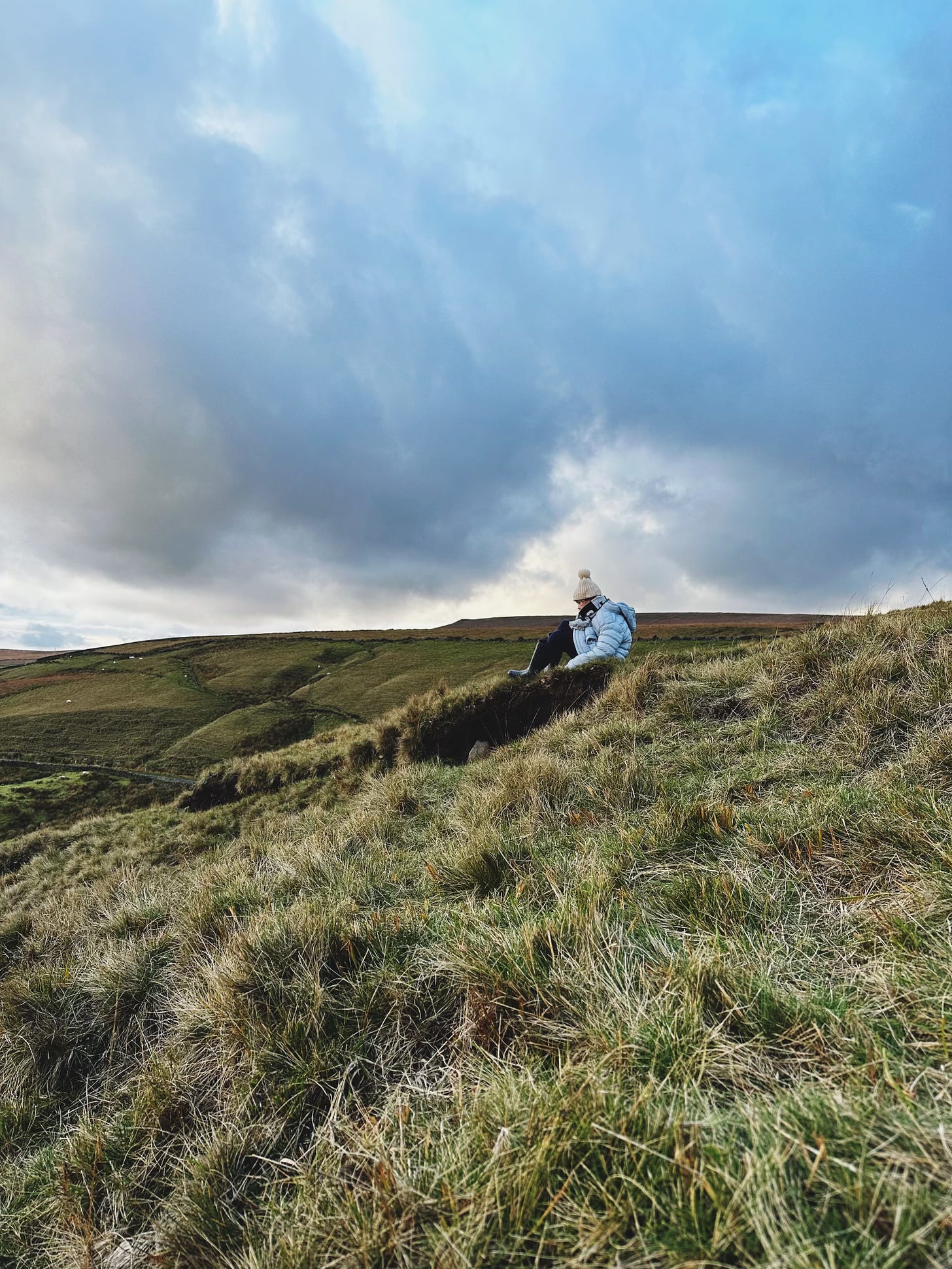 a young girl in a blue coat and white hat sits on a hill in the yorkshire moors. The big sky matches her coat.
