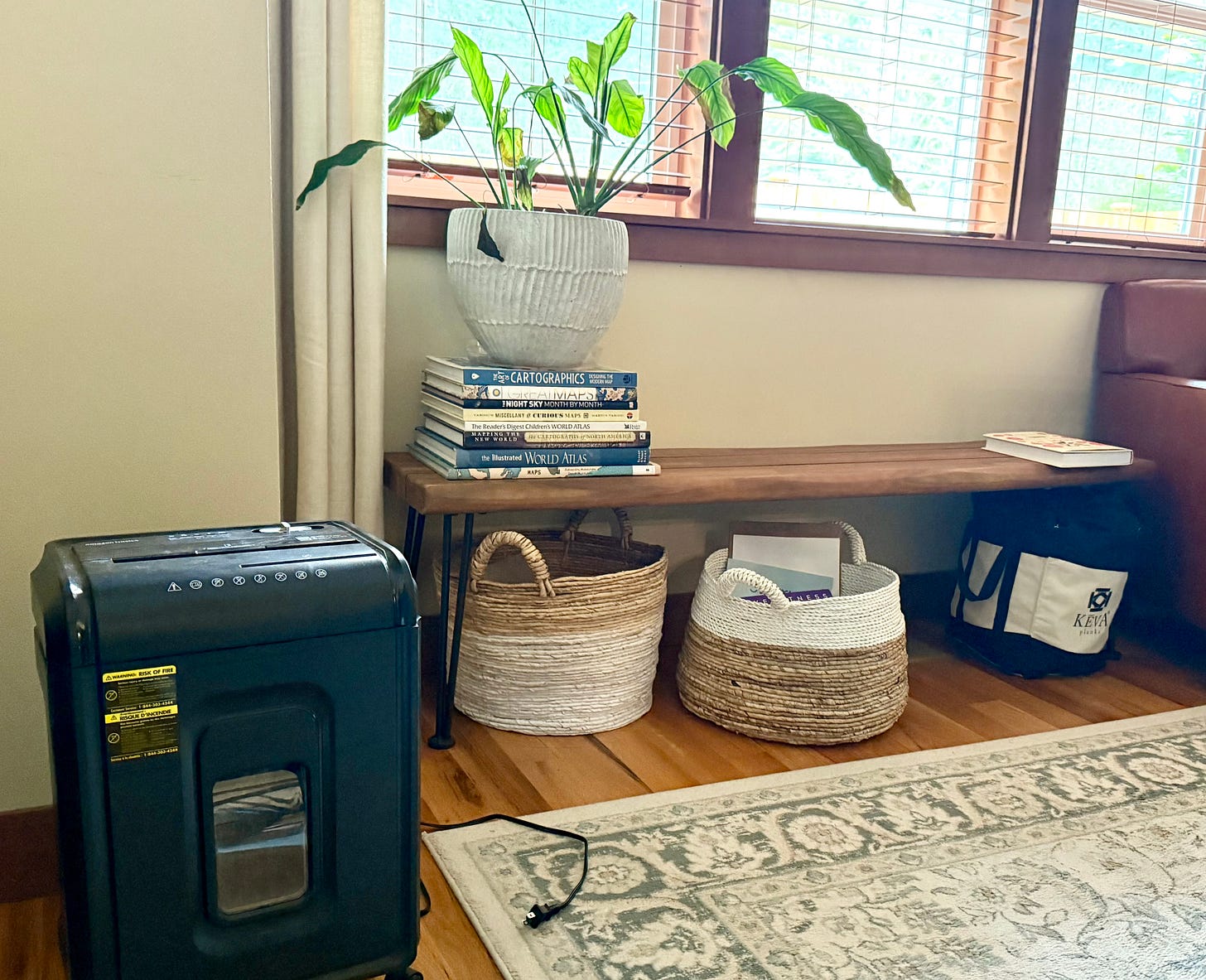 A cozy home corner featuring a simple wooden bench with stacked atlas books and a potted plant, storage baskets underneath, a paper shredder, and natural light coming through wooden blinds on the window