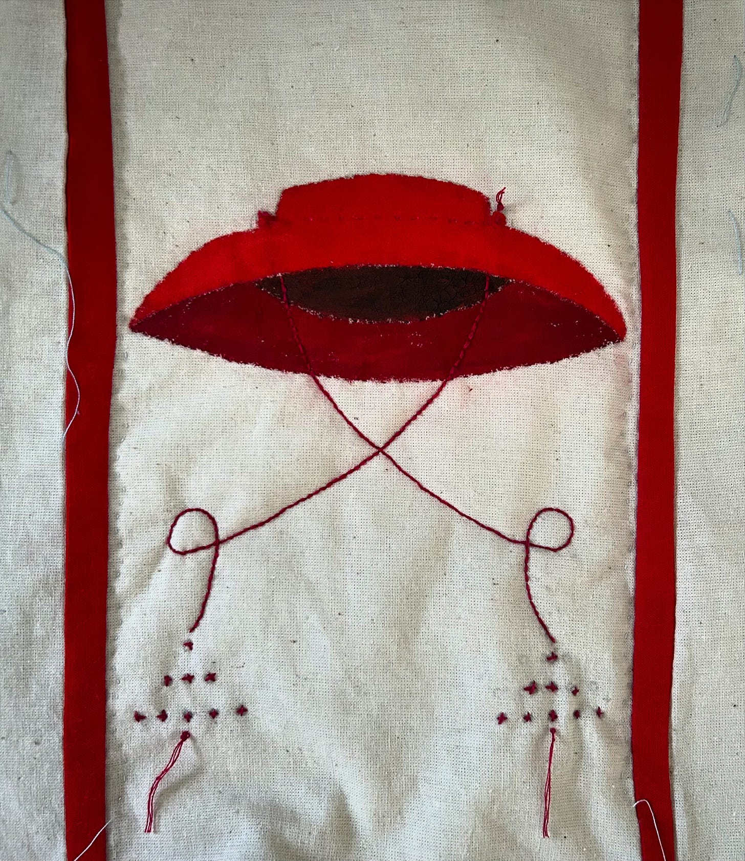 A painted red wide-brimmed cardinal’s hat, with stitched ties, is shown on a piece of cream fabric. Two red strips are shown at each side. 