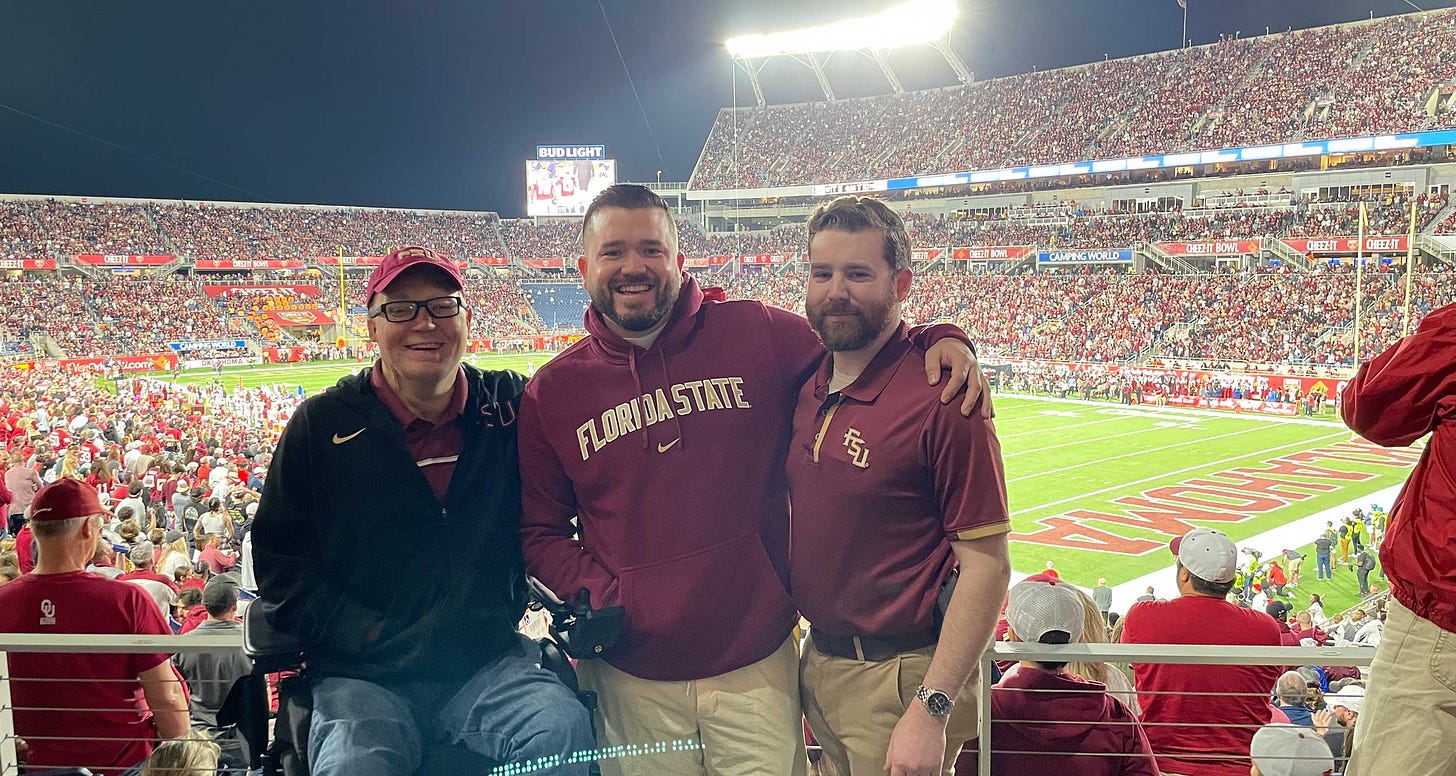 John seated in his wheelchair next to two friends inside a college football stadium.