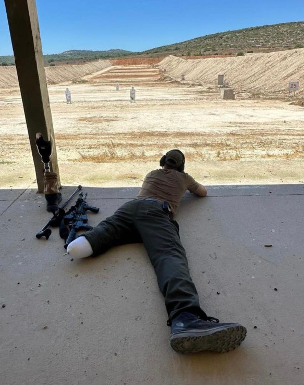 Yoav lies on his stomach with gun aimed into the desert. he is wearing an army uniform. his left leg is amputated below his knee.