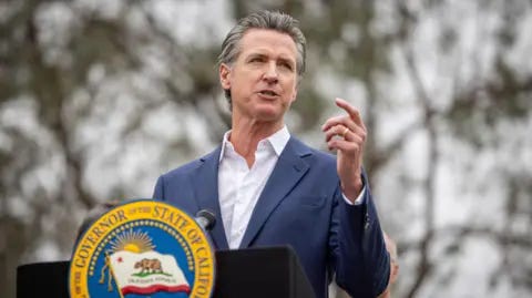 Getty Images California Governor Gavin Newsom standing at a lectern, speaking at a press conference, with one arm slightly raised