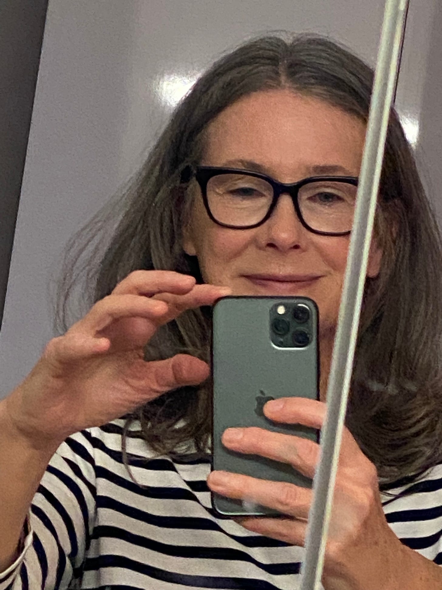 Elizabeth, in a white and blue striped shirt and wearing glasses, takes a bad selfie in the bathroom mirror.