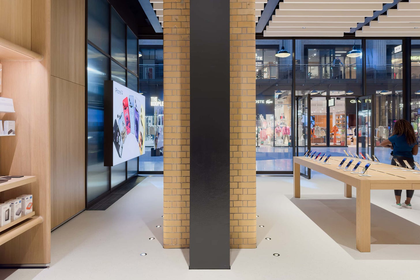 A large steel column sits behind a restored brick column at Apple Battersea. A backlit graphic panel hangs against a glass wall in the distance.