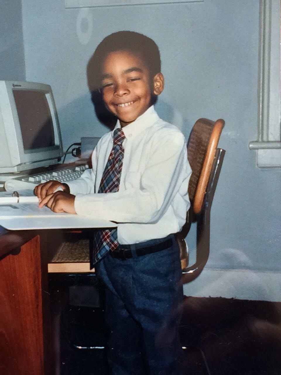 Tomik Dash as a toddler, wearing a white shirt, tie, and dark trousers, with a cheerful smile, sitting at an older computer model in a room with blue walls. Tomik is pointing at a sheet of paper in a white three-ring binder that sits atop a wooden desk.