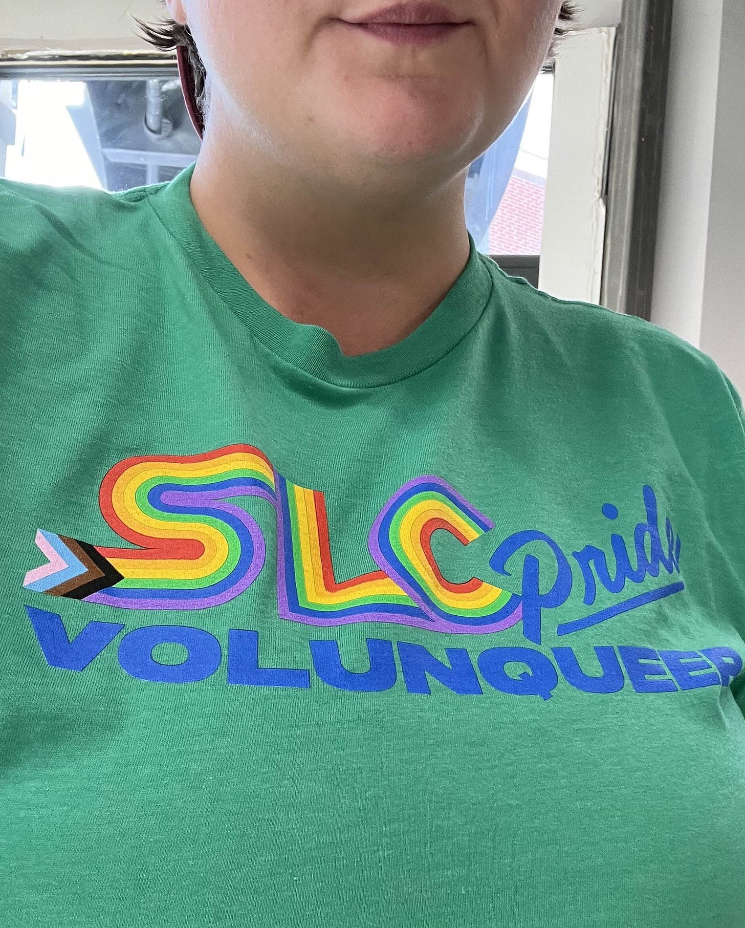 mayson wears a green tee shirt with the slc pride rainbow logo and the word volunqueer in all caps. 