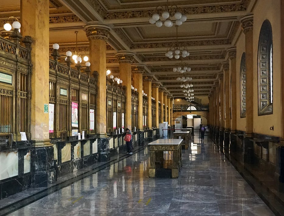 Art Deco Mexico City post office interior hallway. Gold colored marble square pillars line one wall interspersed with brass cages for the tellers. There are long tables in the middle of the hallway and a polished grey marble floor