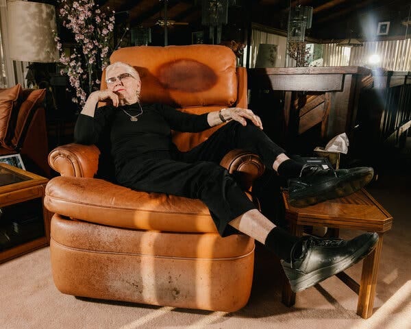Elaine LaLanne reclines on a weathered leather chair wearing all black. Her legs stretch over one arm of the chair as she leans on the opposite arm.
