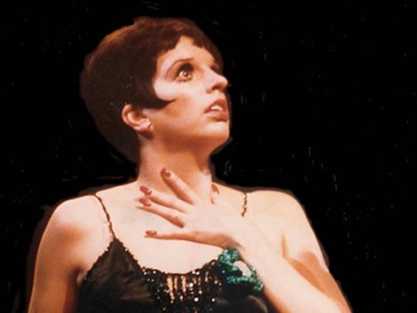 Stage and screen star Liza Minelli, a white woman with short pixie-cut brown hair, clutches her chest, in front of a black background.