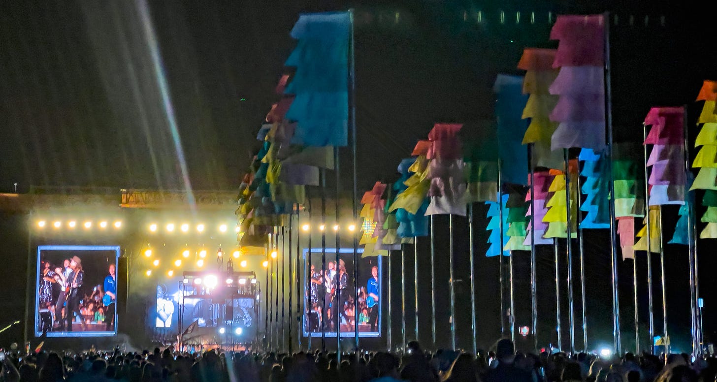 Outdoor stage with large screensm of the musical group on either side of the stage; three rows of colorful flags stand on the right blowing in the wind against the night sky