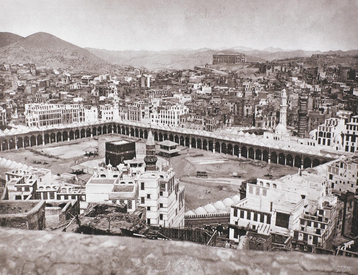 Black and white photo of the al Haram mosque, the Ka'aba at the center of its large rectangular courtyard, and city buildings of Mecca, taken from above on one of the hills surrounding the city. 