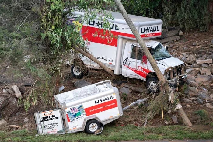 Two U-Haul trucks heavily damaged, crashed against trees and debris, seemingly after a natural disaster like a flood or landslide