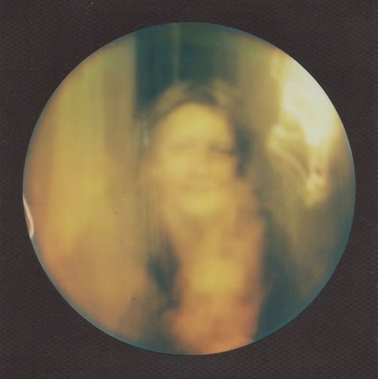 round-shaped polaroid picture in golden hues against a dark background: an out-of-focus person from the torso up next to a light source, wearing a dark sweater and what looks like a flame (and is really a ghost image of the face)