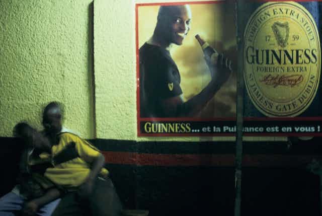 A slightly blurry image of two men hugging playfully, above them a large advert for Guinness beer with a handsome, smiling African man raising a bottle to his lips.