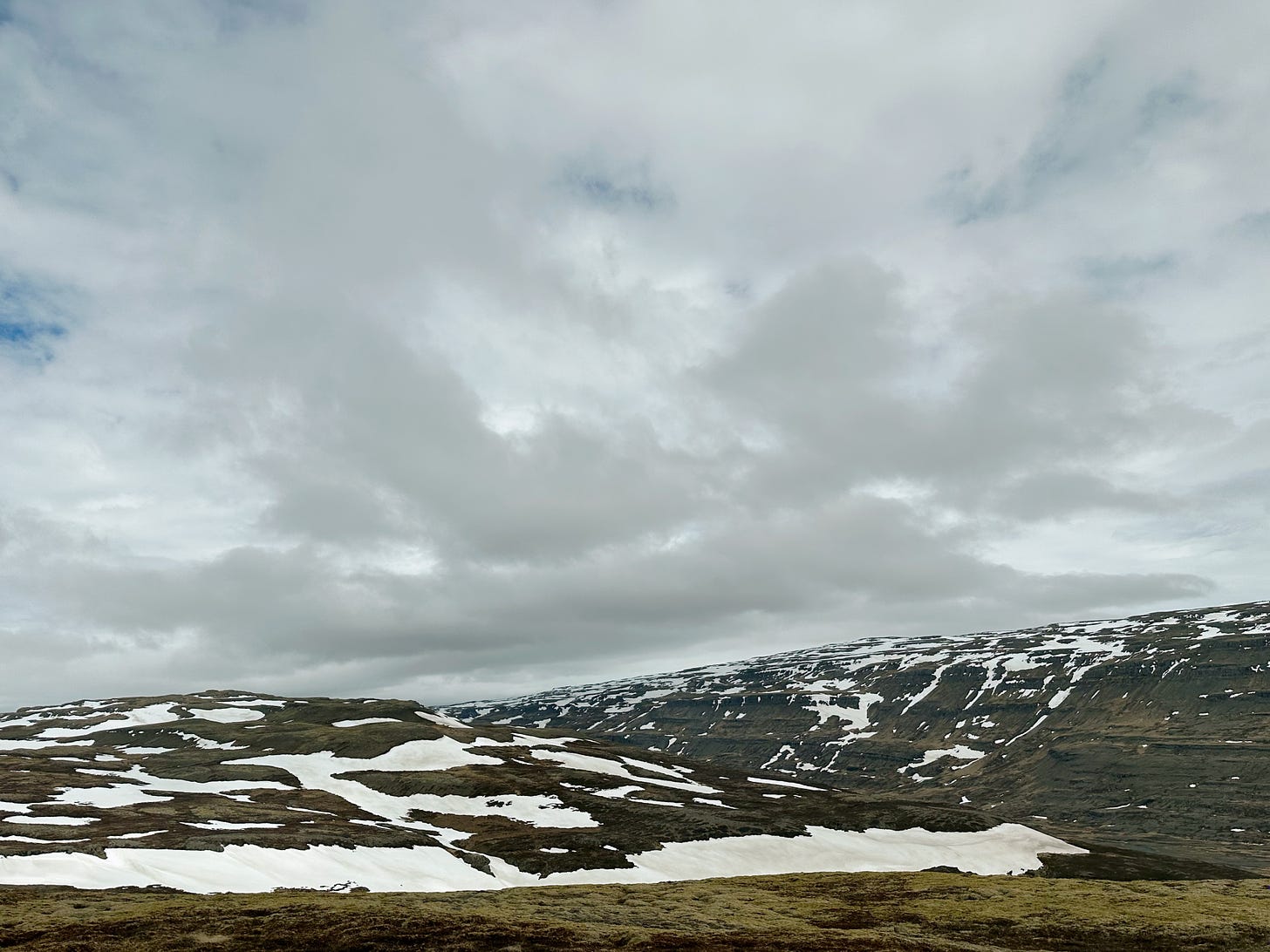 Snow filled vista from the highway.  Snow is just starting to melt in patches along the sides, moss is beginning to green after winter and the sky has blue patches between white and gray clouds.