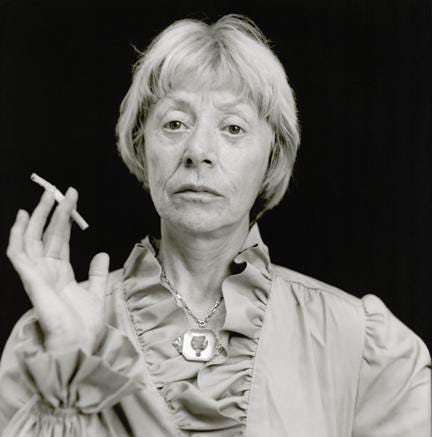 A black-and-white photograph of a light-skinned older woman. She has closed cropped light hair and is shown from the chest up. She wears a long sleeved shirt with a ruffled collar and sleeves and a large silver necklace. In one hand she holds a lit cigarette.