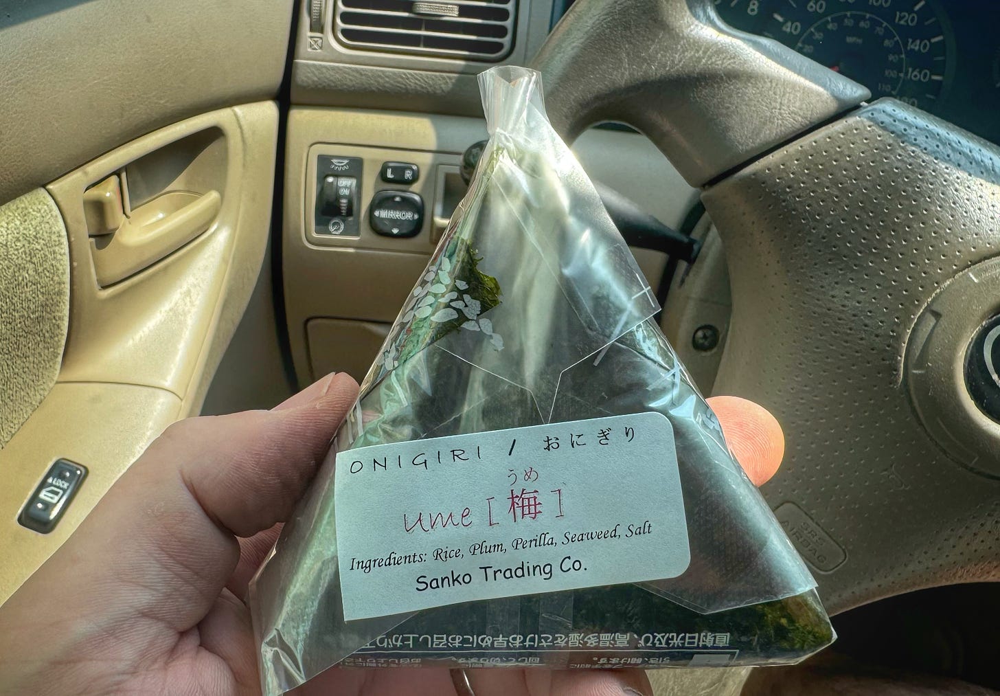 My hand holding a packaged onigiri plum-flavoured rice ball in front of the steering wheel of my car, where I am seated and about to dig in.