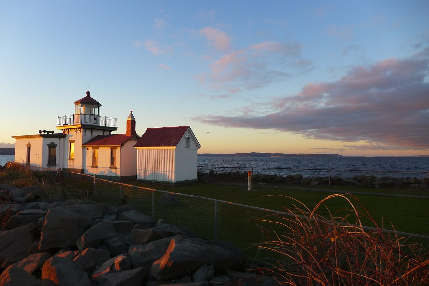 a white lighthouse with a red roof on green grass in front of water while the sun is setting