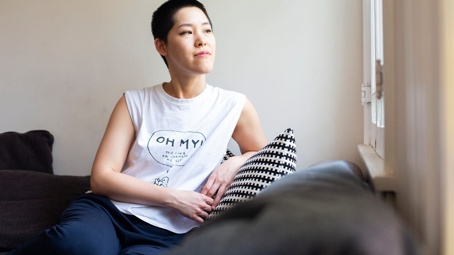 Author photo of Jen Wang looking out a window with a confident smile, her hair buzzed short, wearing a casual white sleeveless tee shirt picturing a cartoon