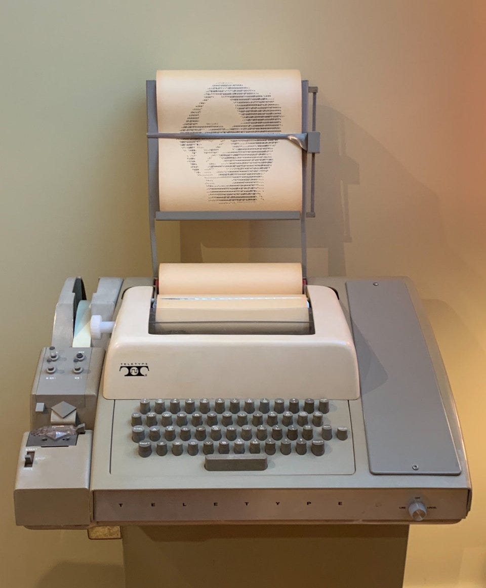 Teletype machine. This picture has some kind of paper holder on the top, with an ASCII-printed picture of somebody I don't recognize. The paper tape reader/writer is on the left. The middle has the keyboard.